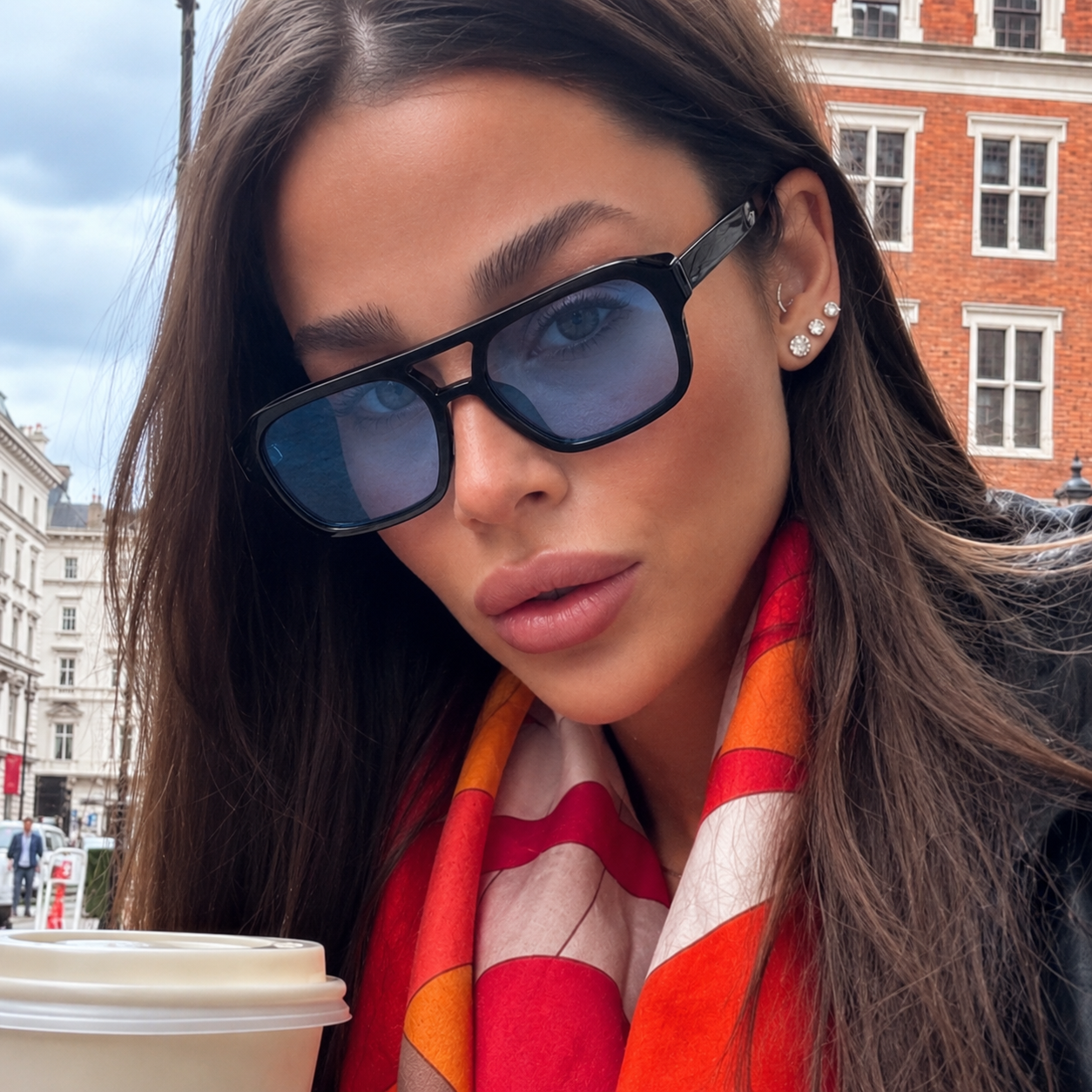 A close-up lifestyle shot of a woman wearing black square sunglasses with blue-tinted lenses, a red, orange, and white patterned scarf, and small pearl stud earrings, set against a blurred background of a city street and brick building.